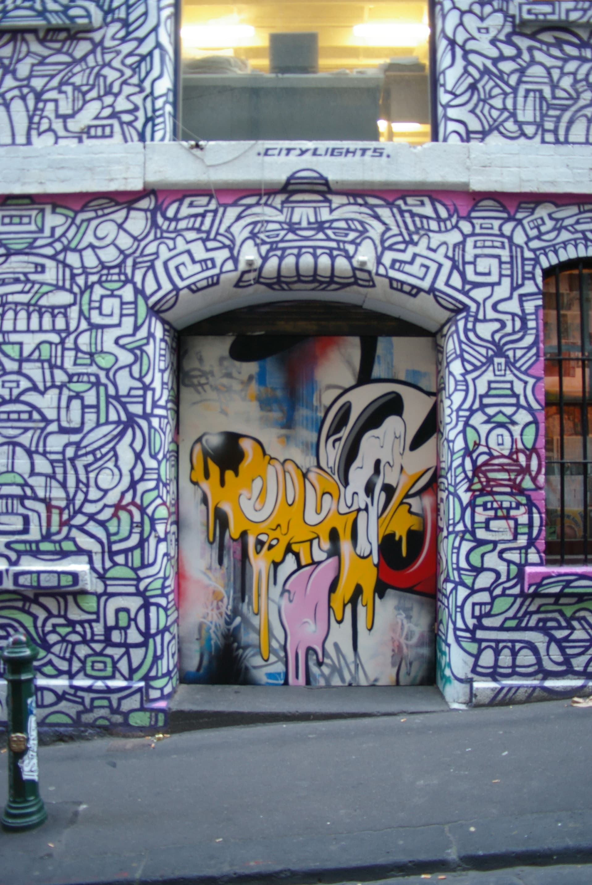 Arched laneway doorway covered in geometric line graffiti, with a dripping cartoon mural on the door and a lit window above