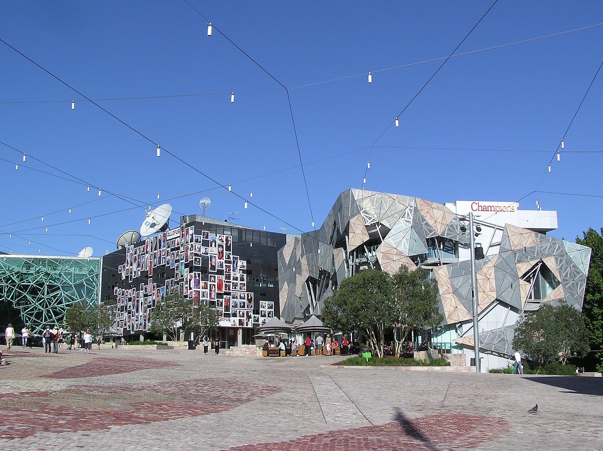 Paved civic plaza with overhead string lights, young trees and faceted contemporary buildings under a clear blue sky
